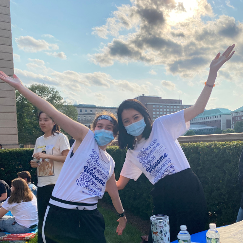 Two students pose with arms outstretched in shirts that say, "welcome"